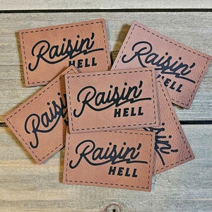 May include: Six brown leather patches with the text "Raisin' Hell" in black lettering. The patches are stitched together and have a rustic, vintage look.