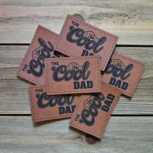 May include: Six brown leather patches with the text "The Cool Dad" in black lettering. The patches have a stitched border and a mountain graphic.