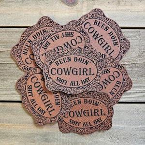 May include: A stack of brown leather coasters with a black floral design and the text "Been doin' Cowgirl Shit all day".