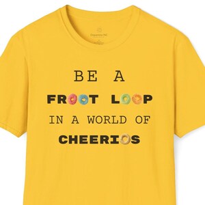 Op de afbeelding: Mosterdgeel t-shirt met de tekst "BE A FROOT LOOP IN A WORLD OF CHEERIOS". De tekst bevat kleurrijke Froot Loops en een Cheerios. Het shirt heeft een ronde hals en korte mouwen.