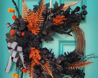 May include: A black and orange Halloween wreath with a black and white striped bow. The wreath is made of faux leaves and flowers and is decorated with black berries and orange pom-poms.
