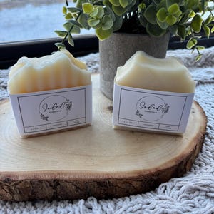 Op de afbeelding: Twee lichtgele zeepblokken, elk met een wit etiket met de tekst "Jaded Essentials" en botanische illustraties. De zepen worden gepresenteerd op een houten schijf, met een plant in een pot op de achtergrond. De zepen wegen ongeveer 113-128 gram.