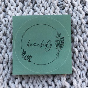 May include: A square, sage green sticker with the word "homebody" in a cursive font, encircled by a black line and floral illustrations. The text "self-proclaimed" is above the circle. The sticker is on a gray, textured surface.