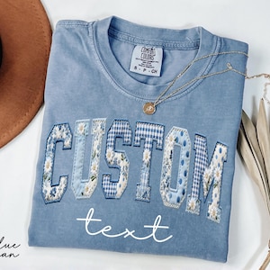 May include: A light blue Comfort Colors t-shirt with the word "CUSTOM" in a patchwork design featuring floral and gingham patterns. A gold necklace and a brown hat are next to the shirt. The word "text" is written below the word "CUSTOM".