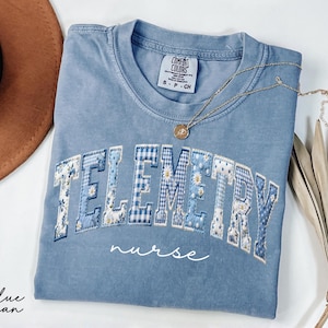 May include: A blue jean Comfort Colors t-shirt with the word "TELEMETRY" in a patchwork design with floral and gingham patterns. The word "nurse" is written in script below. A gold necklace and a brown hat are also in the image.