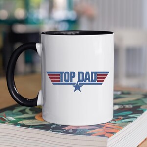 Puede incluir: Taza de cer&aacute;mica blanca con asa e interior negros. La taza presenta el texto "TOP DAD" en un dise&ntilde;o azul, rojo y blanco con una estrella. La taza est&aacute; sobre un libro con un dise&ntilde;o floral.