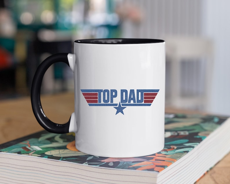 Puede incluir: Taza de cer&aacute;mica blanca con asa e interior negros. La taza presenta el texto "TOP DAD" en un dise&ntilde;o azul, rojo y blanco con una estrella. La taza est&aacute; sobre un libro con un dise&ntilde;o floral.