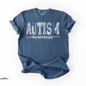 May include: Denim blue t-shirt with the words "AUTISM awareness" in a decorative font. The word "AUTISM" is composed of various patterns, including floral and plaid designs. The shirt is folded, with the sleeves rolled up.