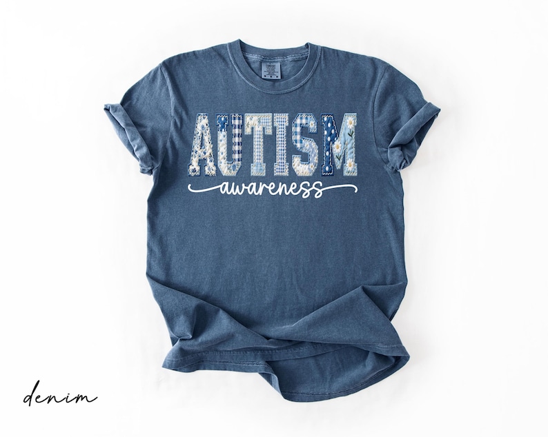May include: Denim blue t-shirt with the words "AUTISM awareness" in a decorative font. The word "AUTISM" is composed of various patterns, including floral and plaid designs. The shirt is folded, with the sleeves rolled up.
