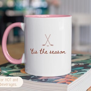 May include: White ceramic mug with a pink interior and handle. The mug features crossed hockey sticks and the phrase "'tis the season" in brown script. The mug is designed for hot and cold drinks.