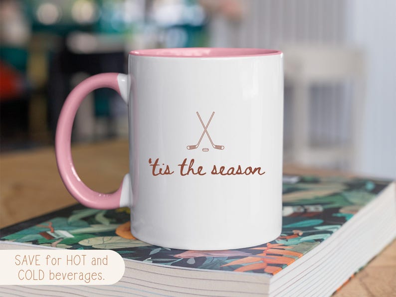 May include: White ceramic mug with a pink interior and handle. The mug features crossed hockey sticks and the phrase "'tis the season" in brown script. The mug is designed for hot and cold drinks.