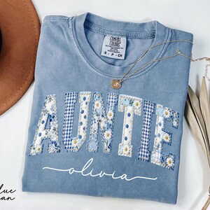 May include: A blue jean Comfort Colors t-shirt with the word "AUNTIE" in a patchwork design of floral and gingham patterns. The name "olivia" is written in cursive below the word "AUNTIE". A gold necklace and a brown hat are also in the image.