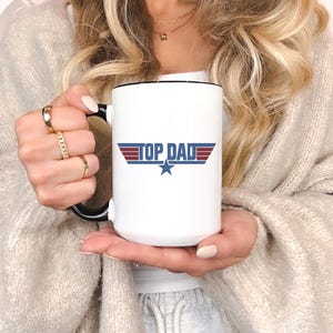 Puede incluir: Taza de cer&aacute;mica blanca con interior y asa negros. La taza presenta el texto "TOP DAD" en azul y rojo, con una estrella azul debajo. Un regalo ideal para el D&iacute;a del Padre.