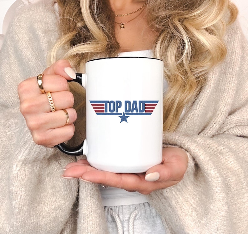 Puede incluir: Taza de cer&aacute;mica blanca con interior y asa negros. La taza presenta el texto "TOP DAD" en azul y rojo, con una estrella azul debajo. Un regalo ideal para el D&iacute;a del Padre.