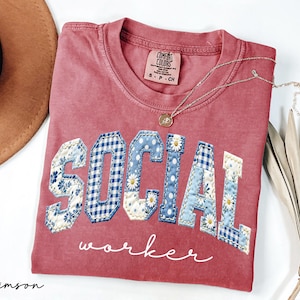 Op de afbeelding: Karmijnrood T-shirt met de woorden "SOCIAL WORKER" in een patchwork design. De letters hebben bloemen-, ruit- en stippenpatronen. Een gouden ketting met hanger ligt op het shirt.