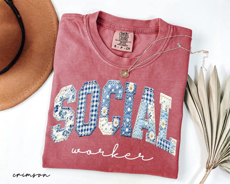 Op de afbeelding: Karmijnrood T-shirt met de woorden "SOCIAL WORKER" in een patchwork design. De letters hebben bloemen-, ruit- en stippenpatronen. Een gouden ketting met hanger ligt op het shirt.