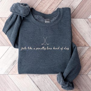 Op de afbeelding: Een marineblauwe sweatshirt met de tekst "feels like a penalty box kind of day" in cursieve letters. Boven de tekst staan twee gekruiste hockeysticks. De sweatshirt ligt op een lichtbruin en wit houten oppervlak.