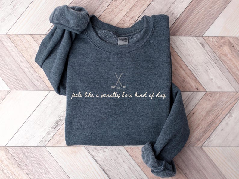 Op de afbeelding: Een marineblauwe sweatshirt met de tekst "feels like a penalty box kind of day" in cursieve letters. Boven de tekst staan twee gekruiste hockeysticks. De sweatshirt ligt op een lichtbruin en wit houten oppervlak.