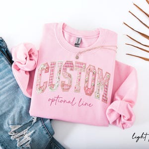 May include: A light pink crewneck sweatshirt featuring the word CUSTOM in large, multi-patterned fabric letters with floral and gingham prints. Below the word, the text optional line is printed in a cursive font. A pair of blue denim jeans is visible.