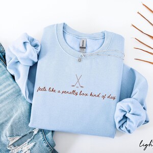 Op de afbeelding: Lichtblauw sweatshirt met de tekst "feels like a penalty box kind of day" en een hockeystick-afbeelding. Een zilveren ketting ligt op het sweatshirt. Het sweatshirt wordt gecombineerd met een blauwe jeans.