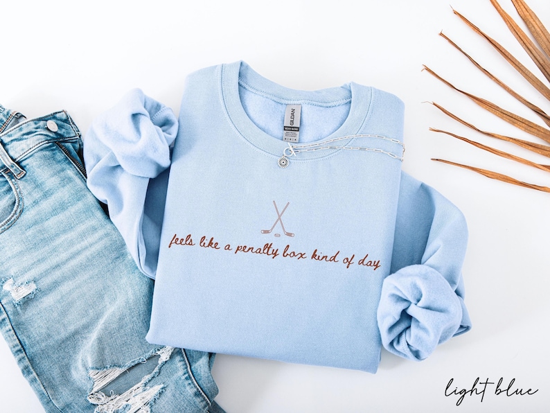 Op de afbeelding: Lichtblauw sweatshirt met de tekst "feels like a penalty box kind of day" en een hockeystick-afbeelding. Een zilveren ketting ligt op het sweatshirt. Het sweatshirt wordt gecombineerd met een blauwe jeans.