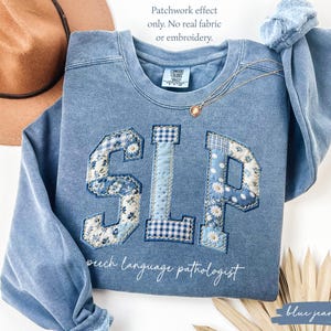 May include: A blue crewneck sweatshirt featuring the letters SLP in a printed patchwork design with floral, gingham, and polka dot patterns. Below the letters, the text reads speech language pathologist. A note states: Patchwork effect only. No real fabric or embroidery.
