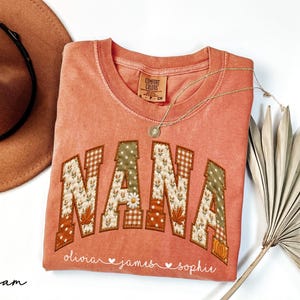 May include: A terracotta-colored t-shirt with the word NANA in large, patchwork-style letters featuring floral, leaf, and gingham patterns. Below the text, the names Olivia, James, and Sophie are printed in white script with small heart icons.