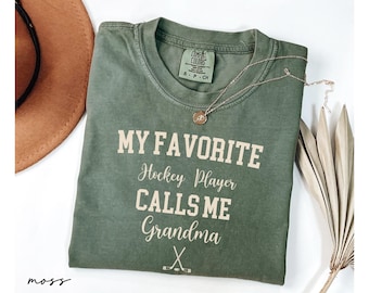 May include: Moss green t-shirt with the text "MY FAVORITE Hockey Player CALLS ME Grandma" in white. A gold necklace and a brown hat are also in the image. The shirt has a Comfort Colors tag.