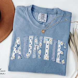 May include: A blue jean Comfort Colors t-shirt with the word "AUNTIE" appliqued across the front in a floral and gingham pattern. A gold necklace with a pendant rests on the shirt. A brown hat is visible in the background.