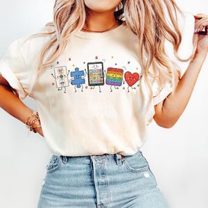 May include: A cream-colored short-sleeve t-shirt featuring a graphic print of five anthropomorphic characters: a white communication board, a blue puzzle piece, a black tablet, a rainbow-colored square fidget toy, and a red heart, all with faces.