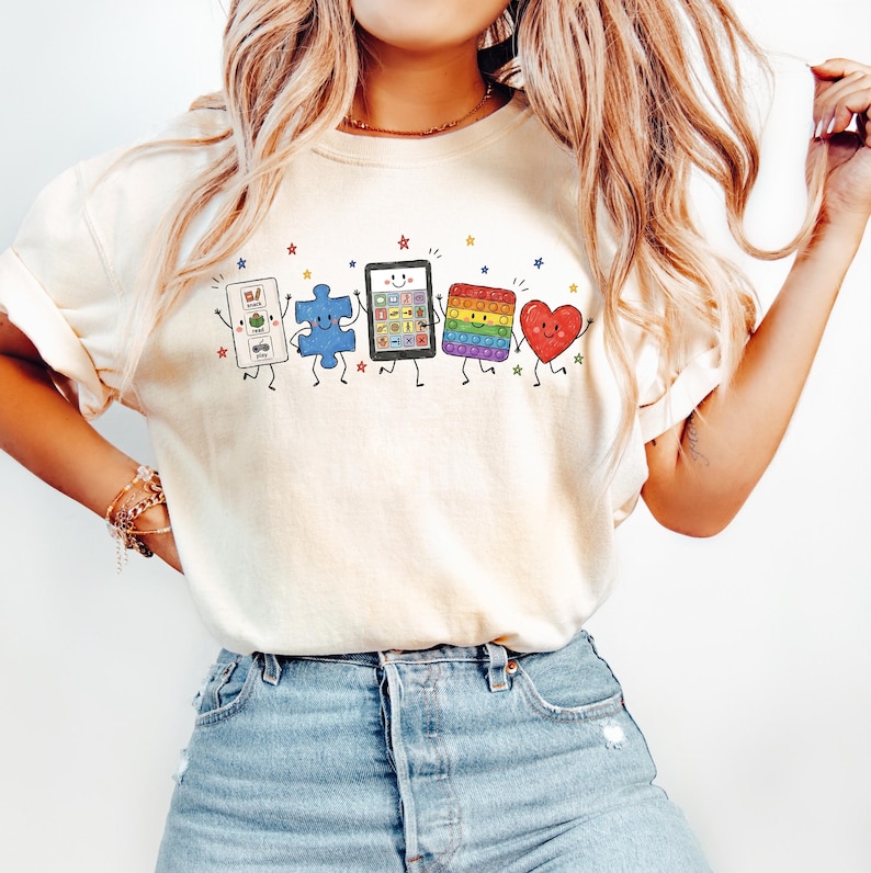 May include: A cream-colored short-sleeve t-shirt featuring a graphic print of five anthropomorphic characters: a white communication board, a blue puzzle piece, a black tablet, a rainbow-colored square fidget toy, and a red heart, all with faces.