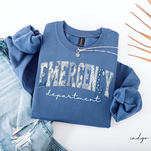 May include: Indigo blue sweatshirt with the words "EMERGENCY department" in a patchwork design. A silver necklace rests on the shirt. The sweatshirt is paired with light blue jeans.