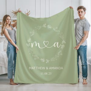 May include: A sage green blanket with a white floral wreath design. The blanket features the initials "m & a" with a heart, and the names "Matthew & Amanda" with the date "11.08.25" in white text. The blanket is held by two people.