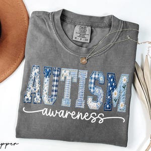 May include: A gray Comfort Colors t-shirt with the word "AUTISM" in blue and white patchwork letters, and the word "awareness" in cursive. A gold necklace and a brown hat are also visible.