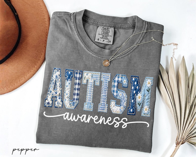 May include: A gray Comfort Colors t-shirt with the word "AUTISM" in blue and white patchwork letters, and the word "awareness" in cursive. A gold necklace and a brown hat are also visible.