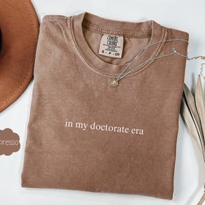 Może przedstawiać: Brązowy t-shirt z nadrukiem "in my doctorate era" w kolorze białym. Koszulka wykonana jest z miękkiego, wygodnego materiału i ma luźny kr&oacute;j.