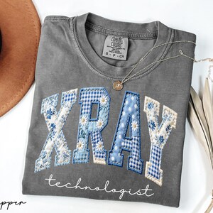 May include: A gray Comfort Colors t-shirt with the word "XRAY" in blue and white patchwork letters, with the word "technologist" below. A gold necklace and a brown hat are next to the shirt.