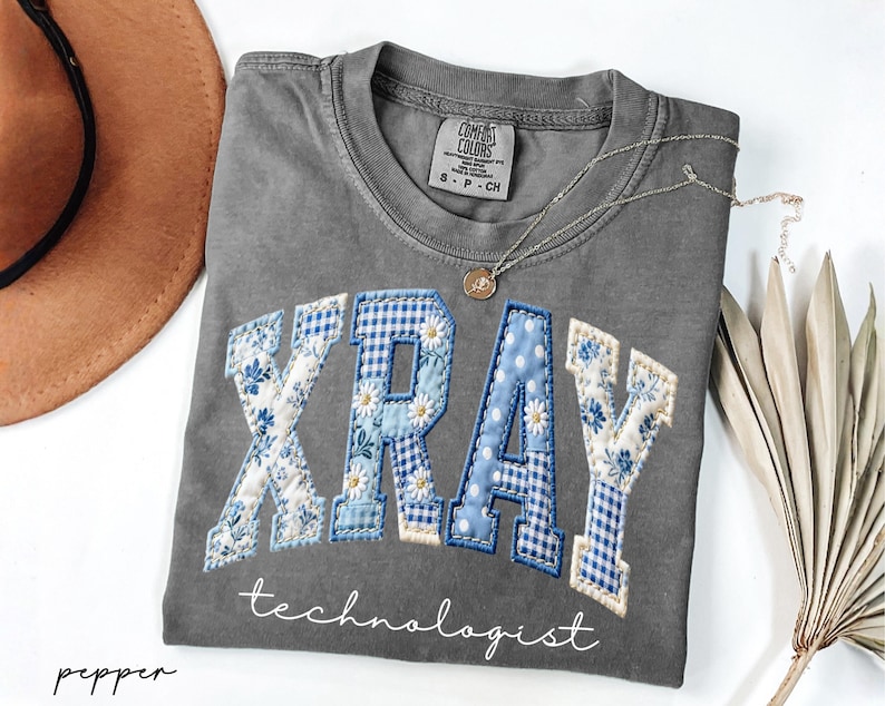 May include: A gray Comfort Colors t-shirt with the word "XRAY" in blue and white patchwork letters, with the word "technologist" below. A gold necklace and a brown hat are next to the shirt.