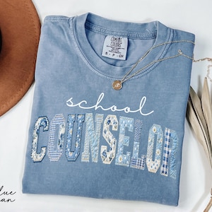 May include: A blue jean Comfort Colors t-shirt with the words "school COUNSELOR" in white and patchwork letters. A gold necklace with a pendant rests on the shirt. A brown hat is in the background.