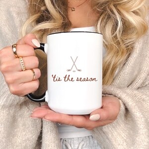 May include: White ceramic mug with a black handle and rim, featuring a hockey stick and puck graphic, and the phrase "'tis the season" in brown script. The mug is held by a person.