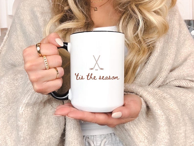 May include: White ceramic mug with a black handle and rim, featuring a hockey stick and puck graphic, and the phrase "'tis the season" in brown script. The mug is held by a person.