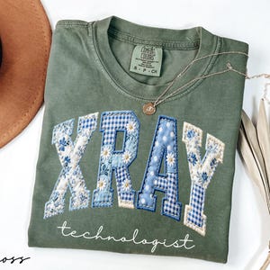 May include: Olive green t-shirt with the word "XRAY" in a patchwork design with blue floral and gingham patterns. The word "technologist" is written below in a script font. A gold necklace with a pendant is draped on the shirt.