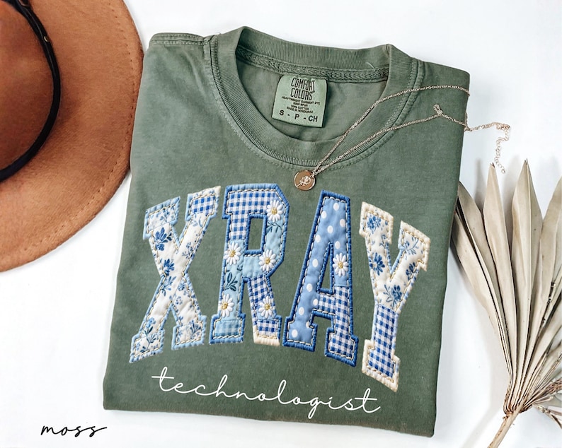 May include: Olive green t-shirt with the word "XRAY" in a patchwork design with blue floral and gingham patterns. The word "technologist" is written below in a script font. A gold necklace with a pendant is draped on the shirt.
