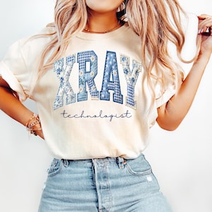 May include: Cream-colored t-shirt featuring the word "XRAY" in blue patchwork letters with floral and gingham patterns. Below the word is the word "technologist" in a cursive font. The shirt is paired with blue denim shorts.