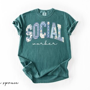 Op de afbeelding: Een tealkleurig t-shirt met de tekst "SOCIAL worker". De letters zijn ontworpen met blauwe en witte bloemen- en ruitpatronen. De woorden "blue spruce" staan in cursiefschrift linksonder.