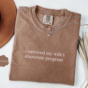 May include: A brown t-shirt with the text "i survived my wife's doctorate program" printed in white. The shirt is folded and laid flat on a surface.