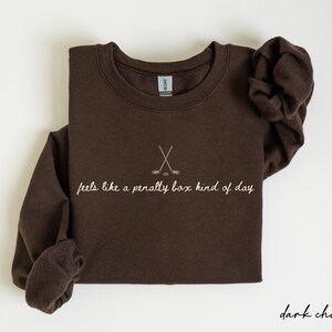 Op de afbeelding: Donker chocoladebruine sweatshirt met een ronde hals en lange mouwen. De voorkant heeft een hockeystick-afbeelding en de zin "feels like a penalty box kind of day" in een cursief lettertype. Het sweatshirt is opgevouwen.