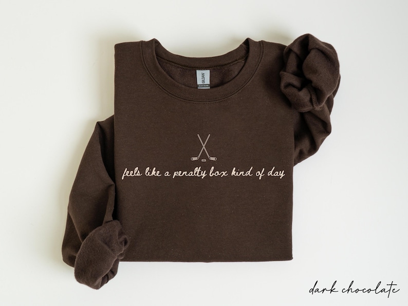 Op de afbeelding: Donker chocoladebruine sweatshirt met een ronde hals en lange mouwen. De voorkant heeft een hockeystick-afbeelding en de zin "feels like a penalty box kind of day" in een cursief lettertype. Het sweatshirt is opgevouwen.