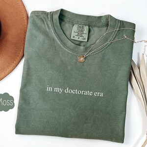 Może przedstawiać: Zielony T-shirt z nadrukiem "in my doctorate era" na przodzie. Koszulka wykonana jest z miękkiego, wygodnego materiału i ma luźny kr&oacute;j.