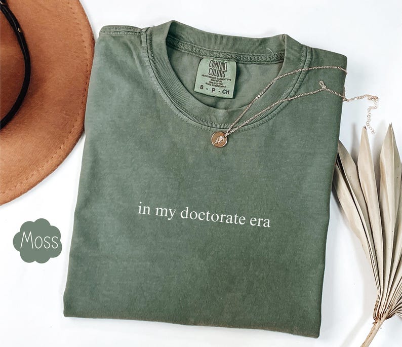 Może przedstawiać: Zielony T-shirt z nadrukiem "in my doctorate era" na przodzie. Koszulka wykonana jest z miękkiego, wygodnego materiału i ma luźny kr&oacute;j.